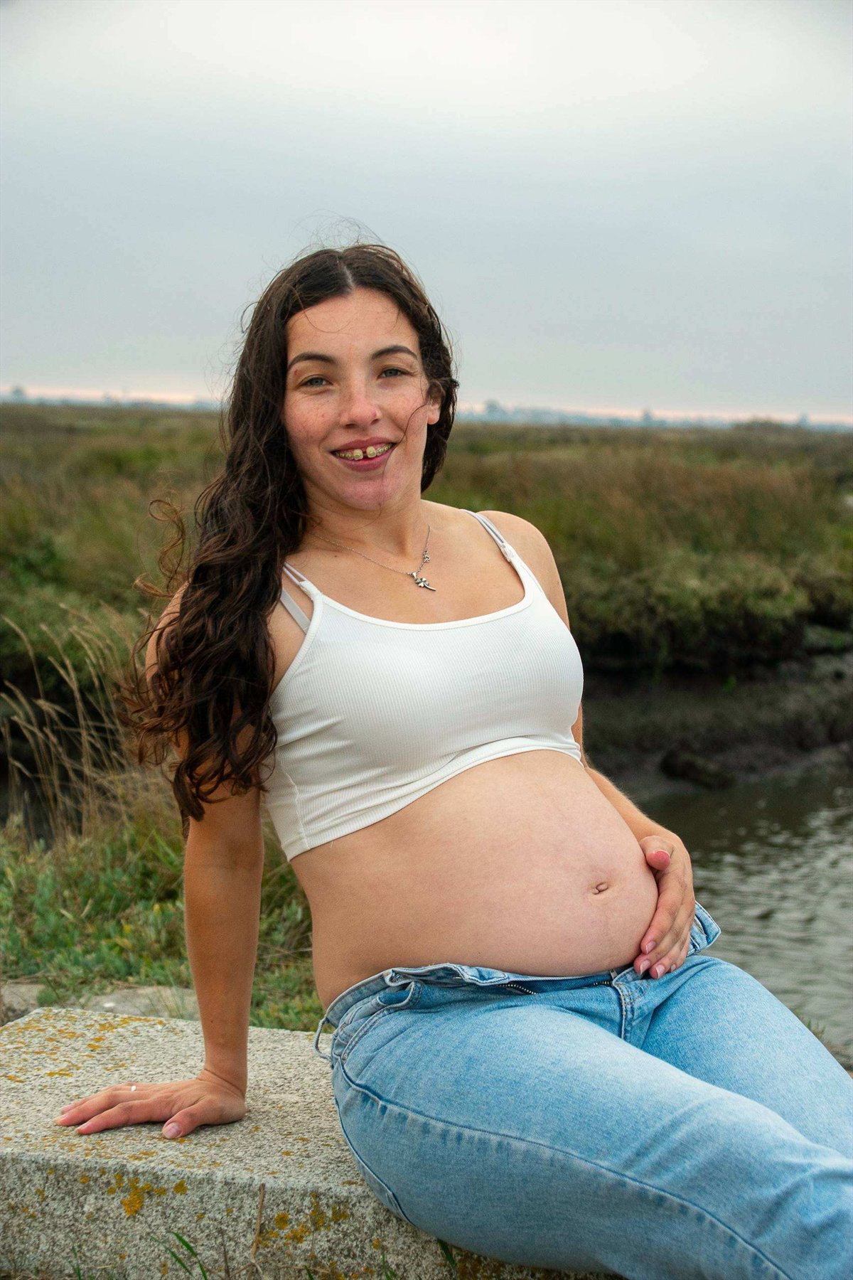 Retrato de grávida com top branco e calças de ganga, sentada num apoio de pedra ao ar livre.
